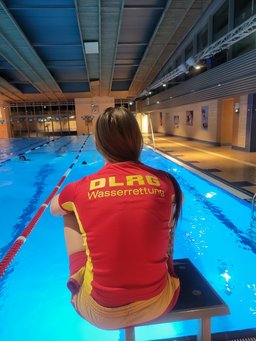 Das Bild zeigt eine Ausbilderin, sitzend auf einem Startblock. Ihr rotes DLRG-Shirt trägt den Schriftzug "Wasserrettung".